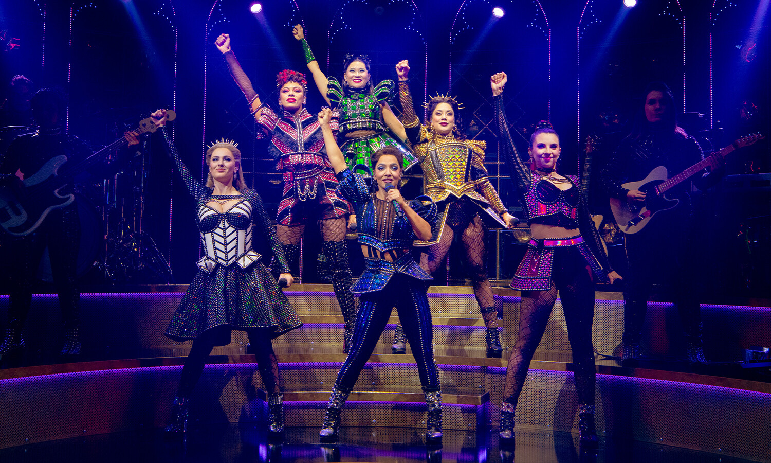 The performers of SIX The Musical stand confidently on stage in vibrant, jewel-encrusted costumes inspired by Tudor fashion. Each queen strikes a powerful pose under dynamic stage lighting, with a backdrop of glowing arches and a live band partially visible. 