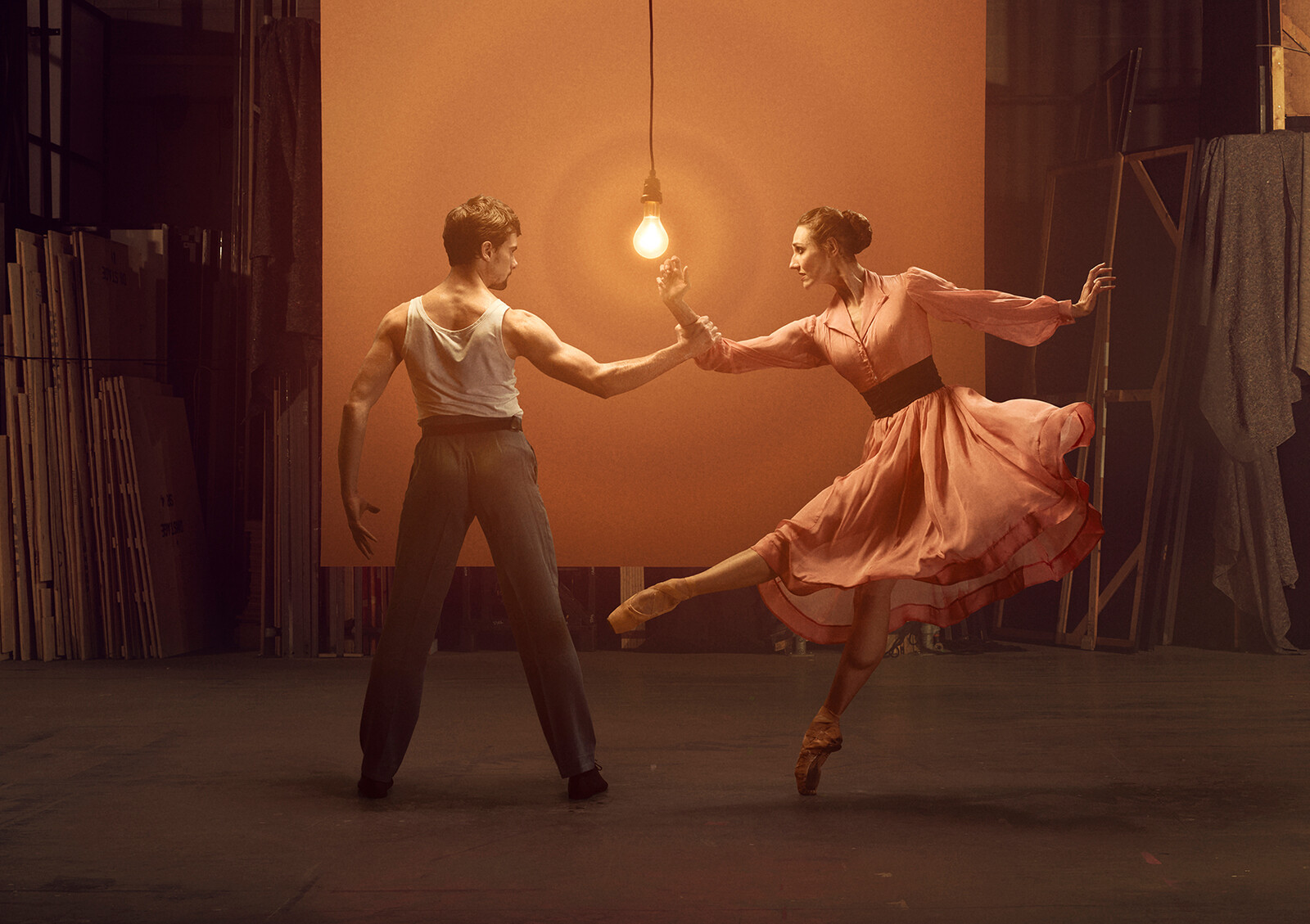 Two dancers perform a dramatic and intimate duet from A Streetcar Named Desire. The man, dressed in a simple tank top and trousers, extends his arm as the woman, elegantly dressed in a flowing pink dress, balances en pointe with graceful precision. A single hanging light bulb casts a warm glow, enhancing the raw and emotional atmosphere of the dimly lit stage