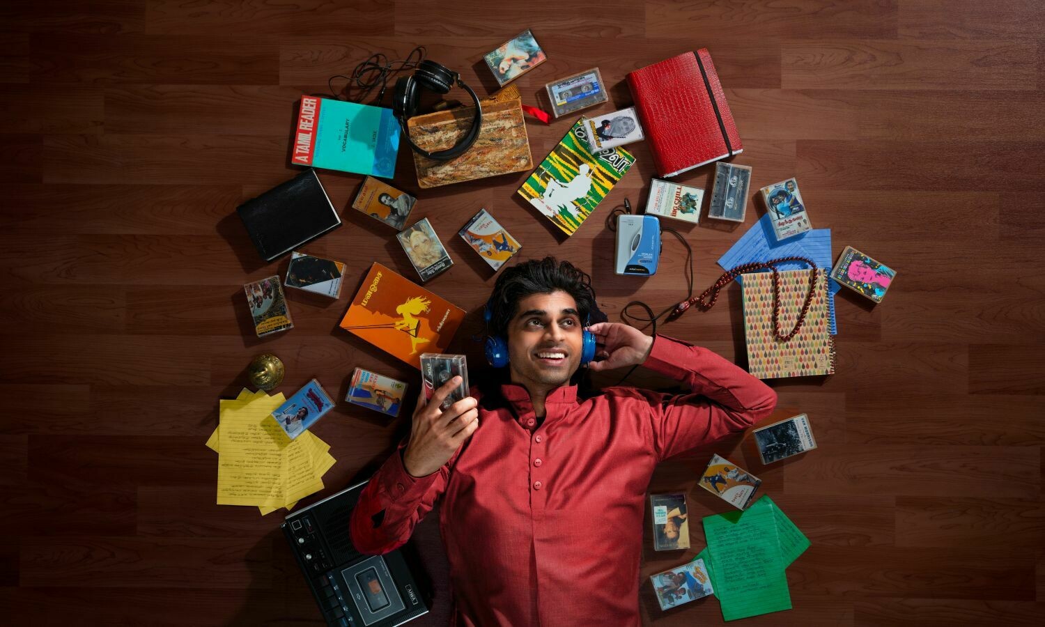 A man in a red kurta lies on a wooden floor, wearing blue headphones and smiling as he holds a cassette tape. Surrounding him are a vibrant assortment of personal items, including books, handwritten notes, cassette tapes, a Walkman, and a tape recorder. The scene evokes a sense of nostalgia and a deep connection to music and storytelling.