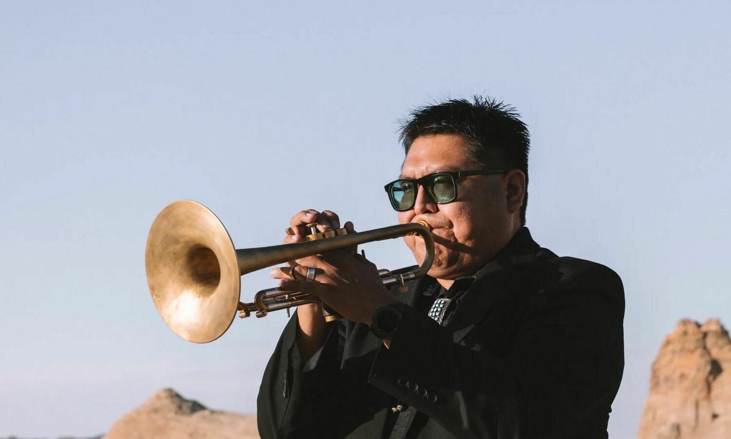 Delbert Anderson, a jazz musician, plays the trumpet outdoors against a serene backdrop of clear skies and desert rock formations. Wearing sunglasses and a sleek black suit, he exudes focus and passion, blending his musical artistry with the natural beauty of the setting.