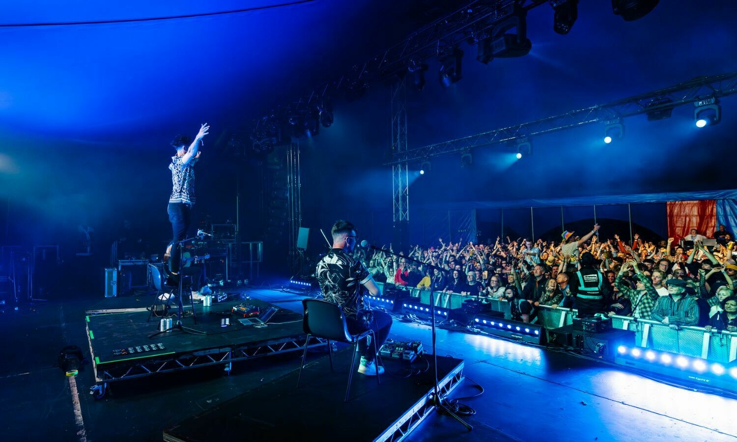 The band Talisk performs live on stage under dynamic blue lighting, captivating a large, enthusiastic crowd. One band member stands on a platform, raising their arms in an energetic gesture, while another sits playing an instrument, facing the audience. The crowd, illuminated by stage lights, cheers and waves their hands, creating a vibrant and electric atmosphere.