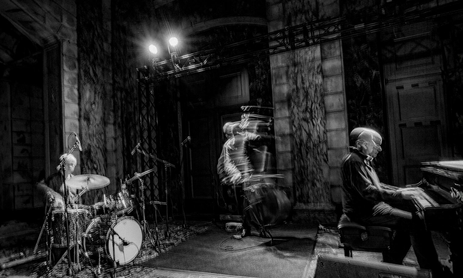 A black-and-white image of the jazz trio The Necks performing on stage. The drummer plays with focused intensity, the pianist leans into the keys with precision, and the bassist's movement is captured in a ghostly blur, conveying dynamic energy. The setting features an ornate architectural backdrop, and the lighting creates a moody, atmospheric feel, emphasising the trio's improvisational style