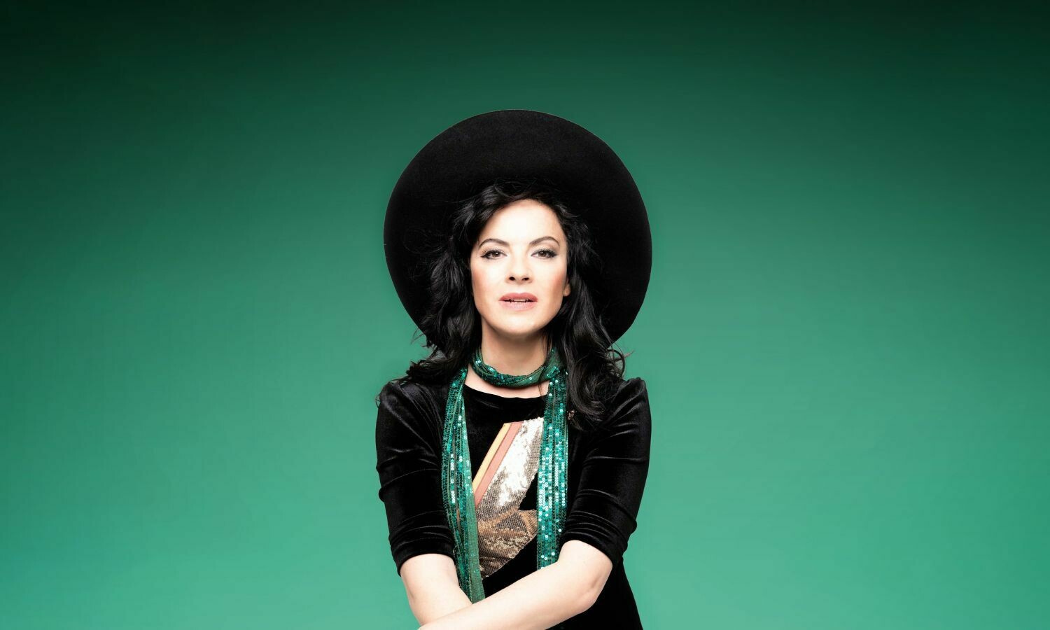 Camille O'Sullivan, the singer, poses against a vibrant green background. She wears a striking black hat and a shimmering green scarf, paired with a black velvet outfit featuring a bold, glittering lightning bolt design. Her confident expression and dramatic styling exude elegance and artistic flair.