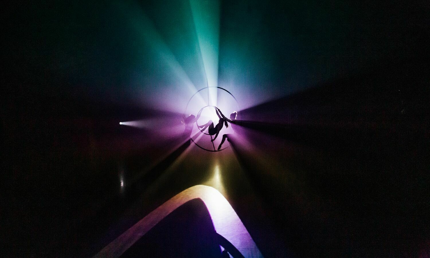 A stunning silhouette of a performer from the show Belle is captured inside a large aerial hoop, suspended in mid-air. Beams of coloured light—purple, green, and gold—radiate outward, creating an ethereal and mesmerising effect. The dark background enhances the dramatic interplay of light and shadow, emphasising the elegance and artistry of the performance.