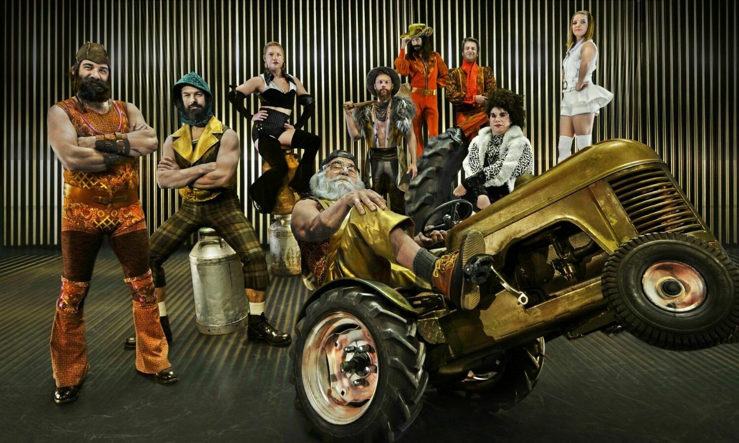 A colourful ensemble of circus performers strikes bold poses against a backdrop of metallic vertical lines. At the centre, a bearded man reclines confidently on a golden tractor, while others surround him in eclectic costumes featuring vibrant patterns, fur, and striking accessories. The group exudes a mix of vintage flair and playful theatricality, creating a lively and eccentric scene.
