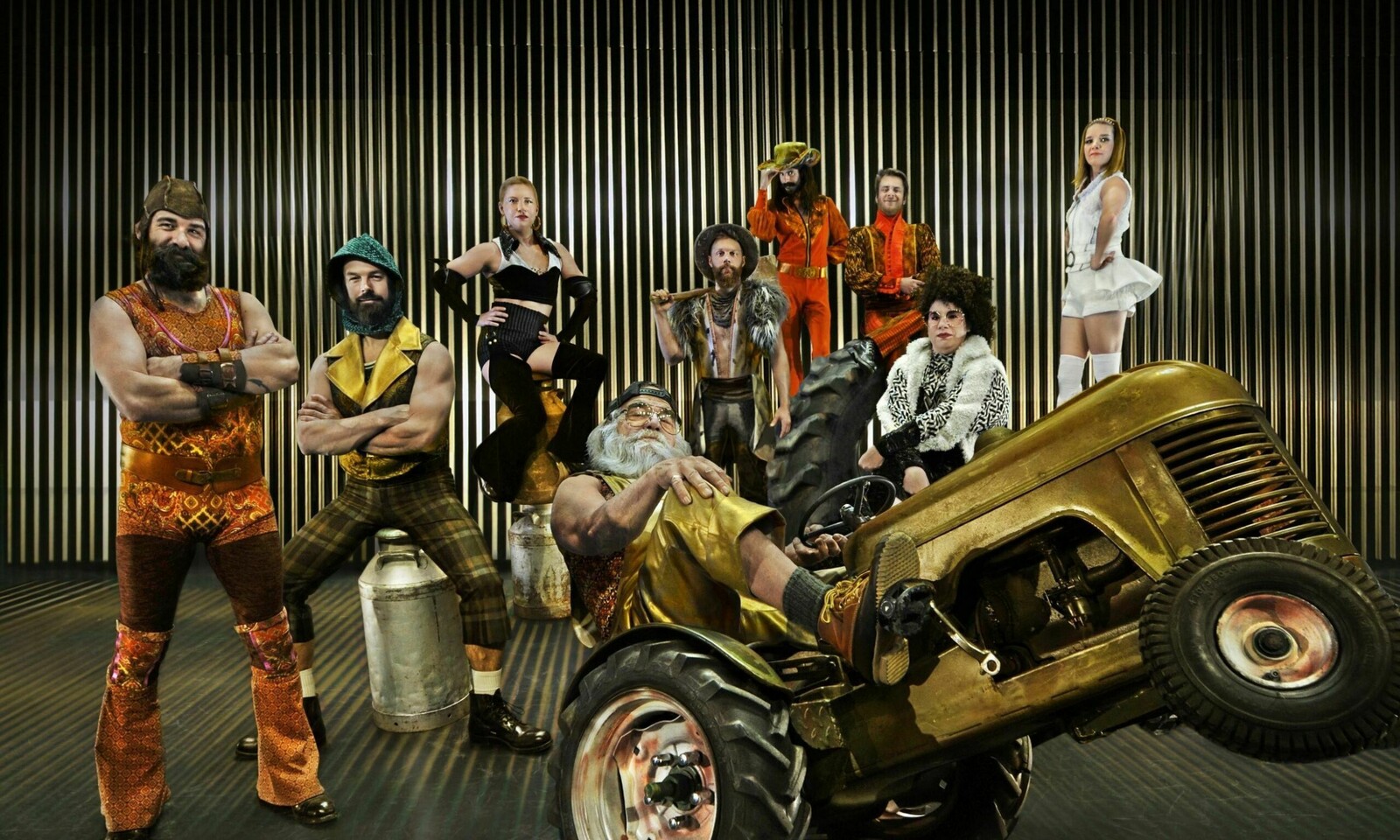 A colourful ensemble of circus performers strikes bold poses against a backdrop of metallic vertical lines. At the centre, a bearded man reclines confidently on a golden tractor, while others surround him in eclectic costumes featuring vibrant patterns, fur, and striking accessories. The group exudes a mix of vintage flair and playful theatricality, creating a lively and eccentric scene.