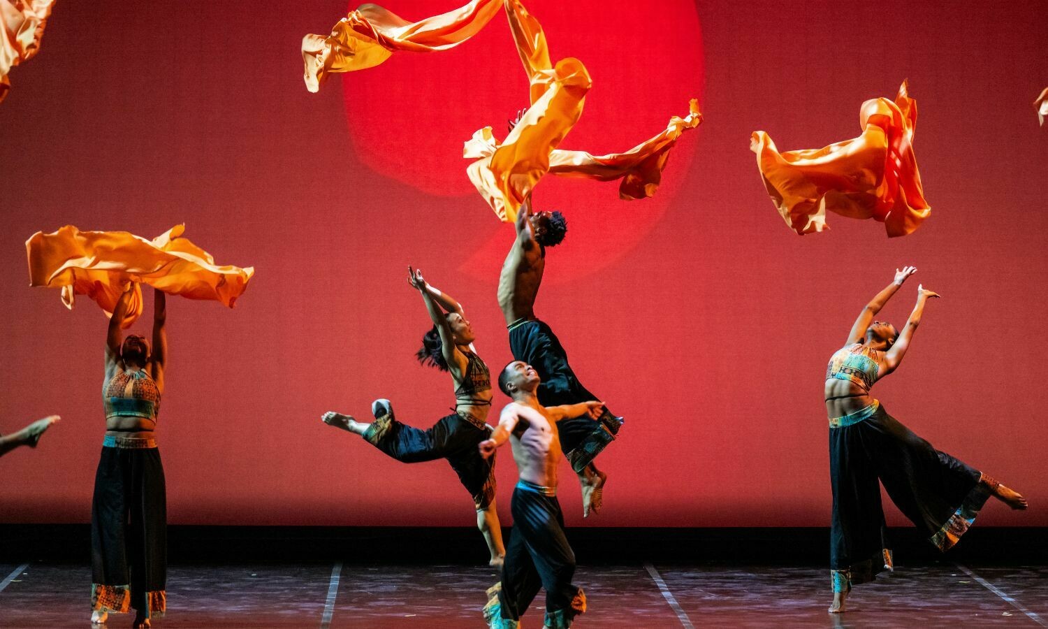 Dancers from the Lula Washington Dance Company perform a captivating routine on stage, their movements synchronised with flowing orange fabric that swirls dramatically through the air. Against a striking red backdrop with a glowing sun-like circle, the performers, dressed in vibrant, patterned costumes, leap and stretch with elegance and power, embodying energy and grace