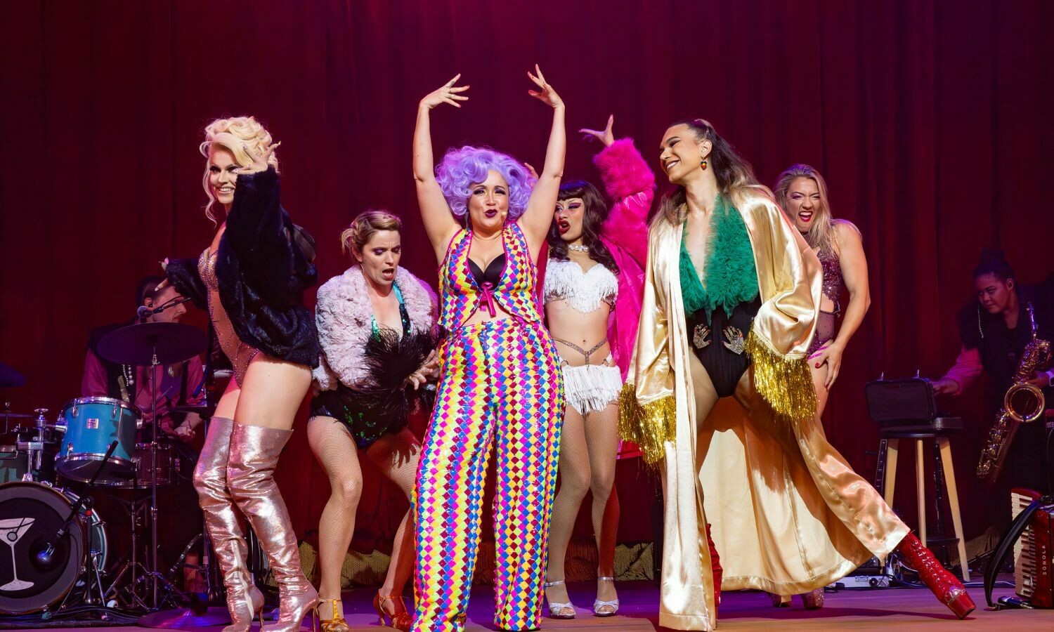 Performers from SMASHED light up the stage in a vibrant scene. Centre stage, a performer with bold purple hair and a colourful harlequin-patterned outfit strikes a dramatic pose, surrounded by cast members in glamorous costumes featuring feathers, sequins, and metallic fabrics. The backdrop of deep red curtains and live musicians in the background adds to the lively, cabaret-style energy of the performance.
