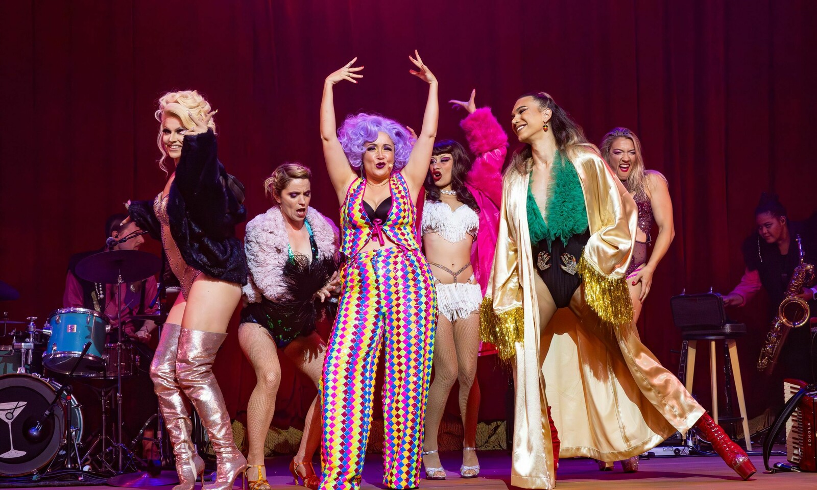 Performers from SMASHED light up the stage in a vibrant scene. Centre stage, a performer with bold purple hair and a colourful harlequin-patterned outfit strikes a dramatic pose, surrounded by cast members in glamorous costumes featuring feathers, sequins, and metallic fabrics. The backdrop of deep red curtains and live musicians in the background adds to the lively, cabaret-style energy of the performance.