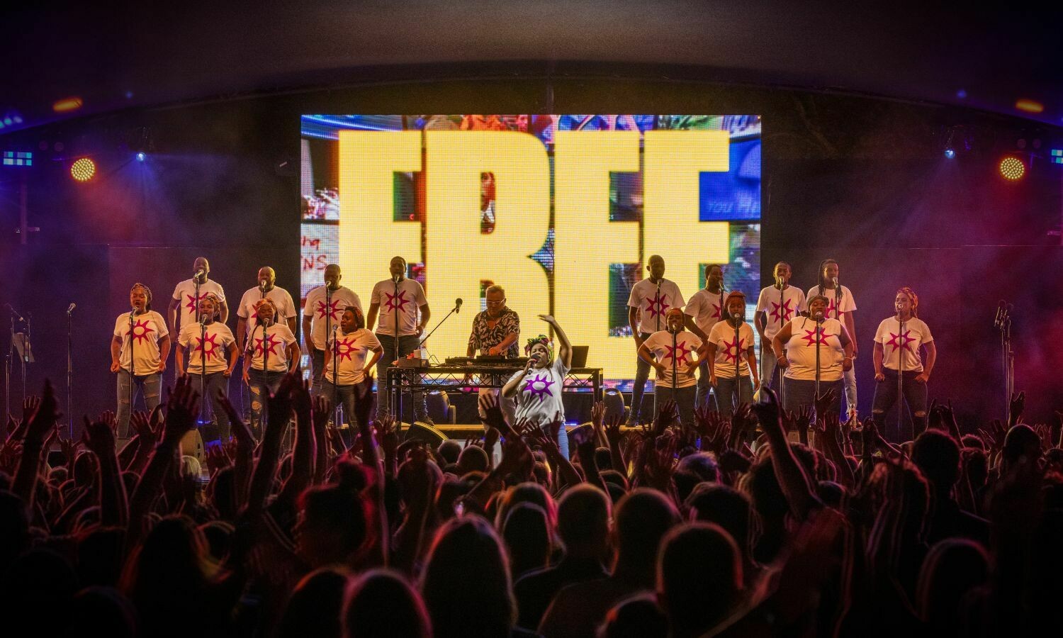 The Soweto Gospel Choir performs energetically on stage alongside DJ Groove Terminator, creating a vibrant and uplifting atmosphere. The choir, dressed in white T-shirts with colourful starburst designs, sings passionately in front of a massive screen displaying the word 'FREE' in bold yellow letters. The stage is bathed in dynamic lighting, with an enthusiastic audience raising their hands in the foreground, fully immersed in the electrifying performance.