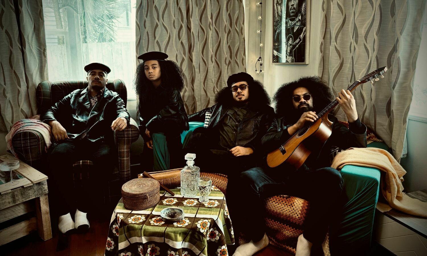 A group of four musicians exudes cool confidence in a cosy, retro-inspired living room setting. Dressed in leather jackets and sunglasses, they sit on mismatched armchairs and sofas, surrounded by vintage decor, including a floral-patterned tablecloth and woven baskets. One member casually plays an acoustic guitar, while the others strike relaxed yet stylish poses. The soft light filtering through the curtains adds warmth to the scene, complementing the laid-back island vibes.