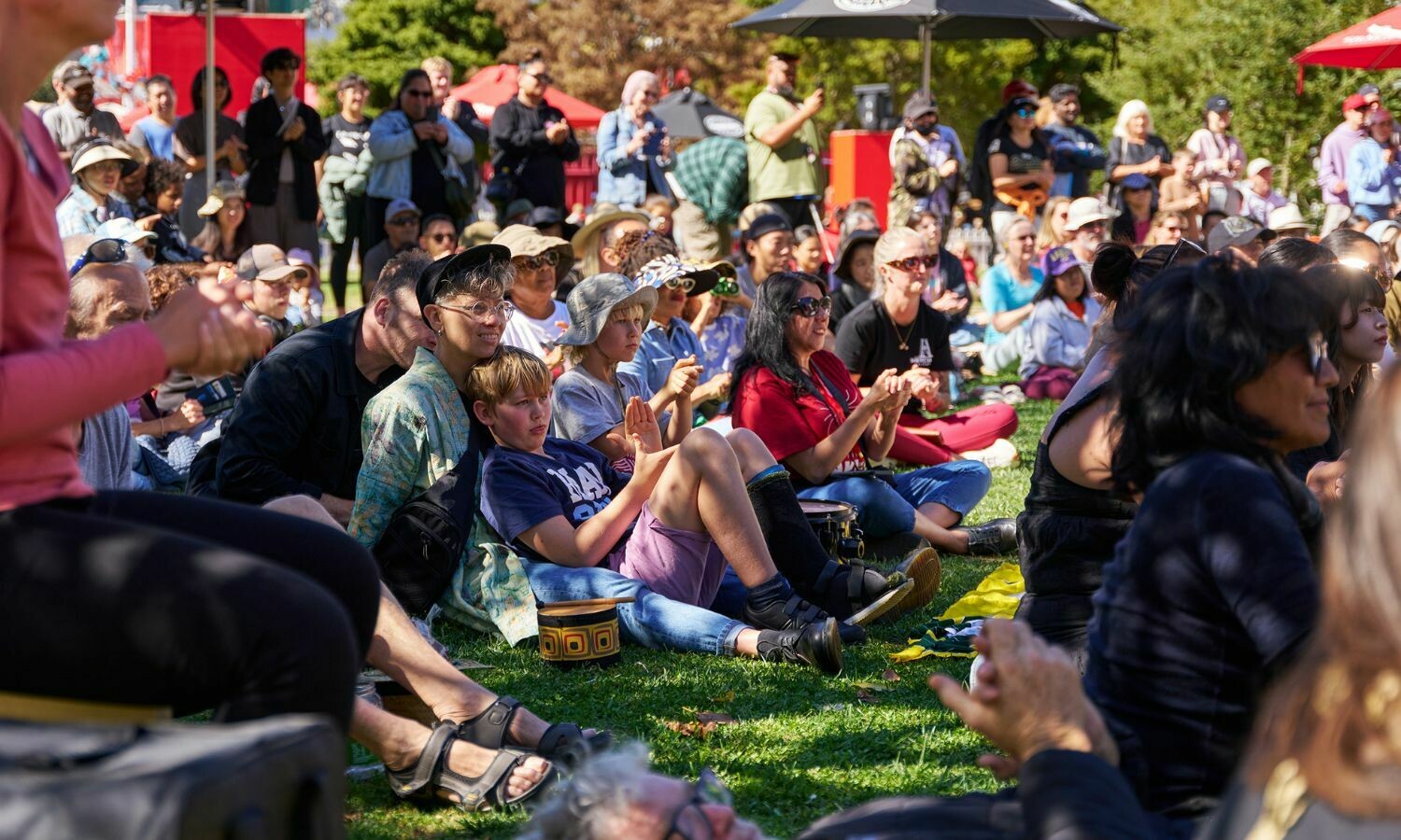 A diverse crowd of families and individuals sits on the grass, enjoying the sunshine during Whānau Day. People of all ages, some wearing hats and sunglasses, clap and smile as they engage with the outdoor event. The relaxed atmosphere is highlighted by vibrant red umbrellas, lush greenery, and a backdrop of spectators standing and capturing the moment on their phones