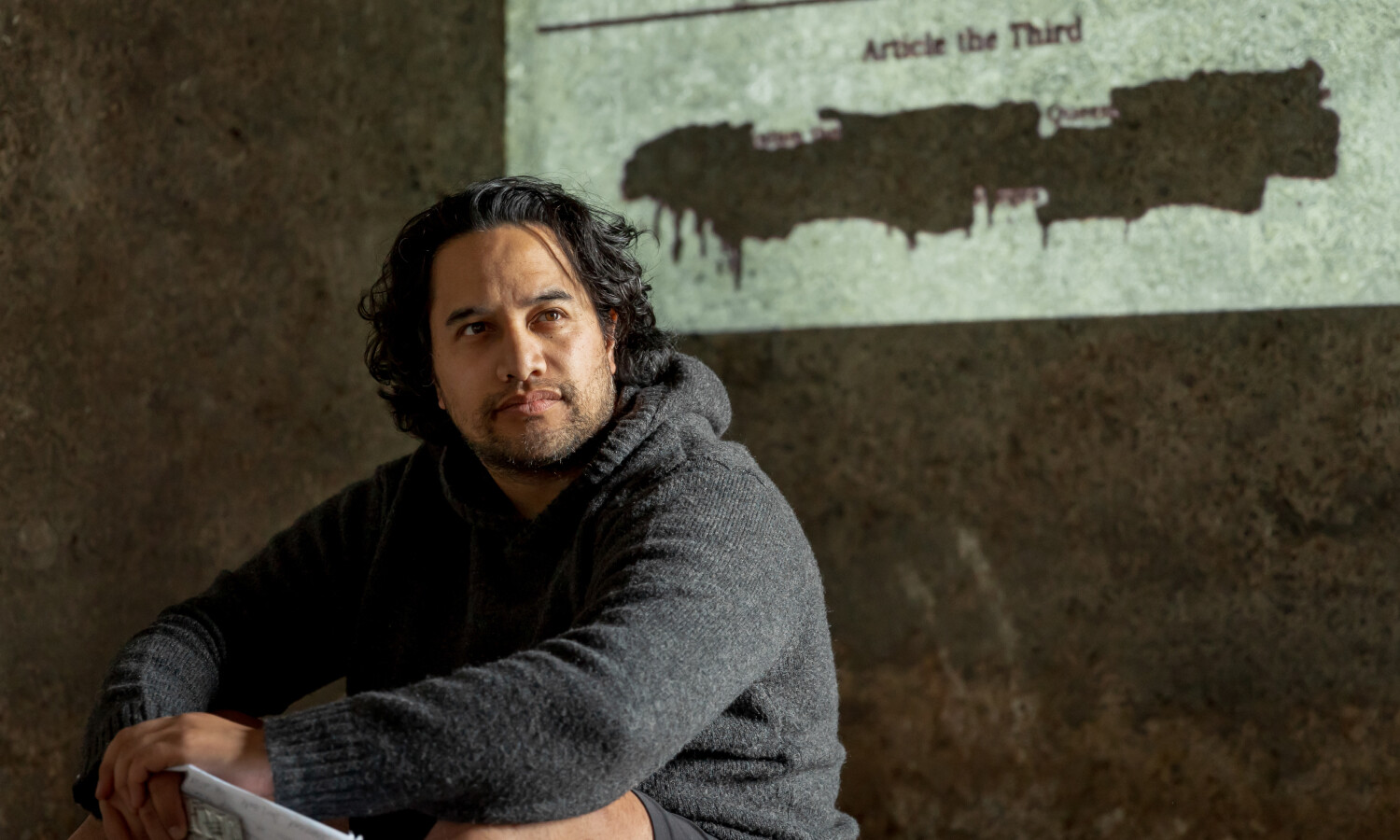 A Māori man sits thoughtfully, dressed in a cosy grey hoodie, with a solemn expression. Behind him, a section of the Treaty of Waitangi is projected onto a textured wall, with parts of the text painted over, symbolising erasure or censorship. The composition evokes themes of history, identity, and reflection.