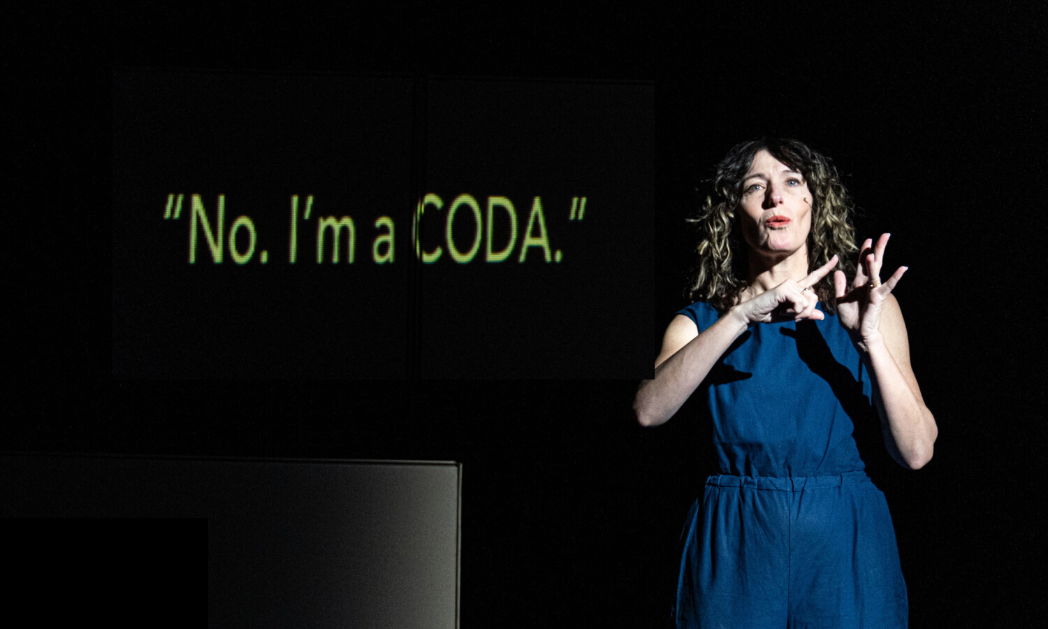 Jodee, a performer in a blue jumpsuit, uses expressive sign language under a focused spotlight. Behind her, the words 'No. I’m a CODA.' are projected in bold yellow text against a black background, emphasising the performance's connection to the CODA (Child of Deaf Adults) experience
